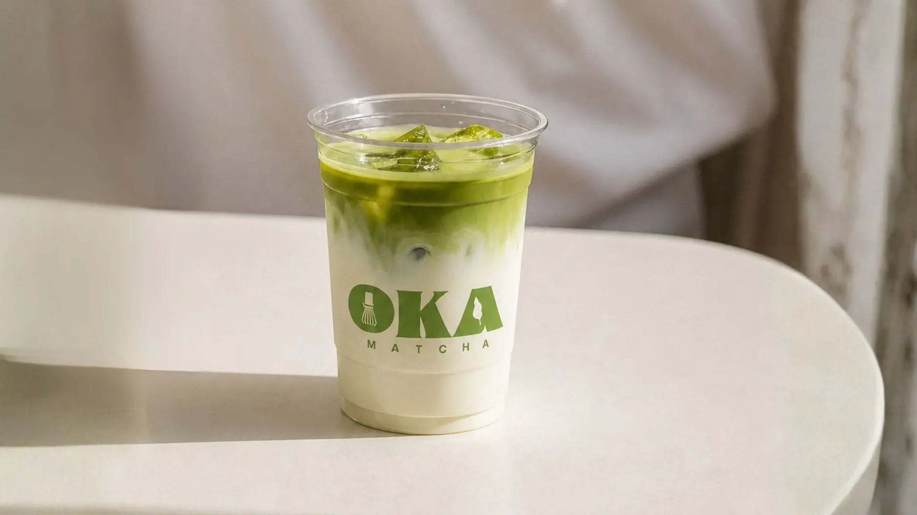 Le gobelet Oka Matcha posé sur une table claire, le latte glacé en dégradé du vert au blanc, le logo imprimé en vert sur la paroi