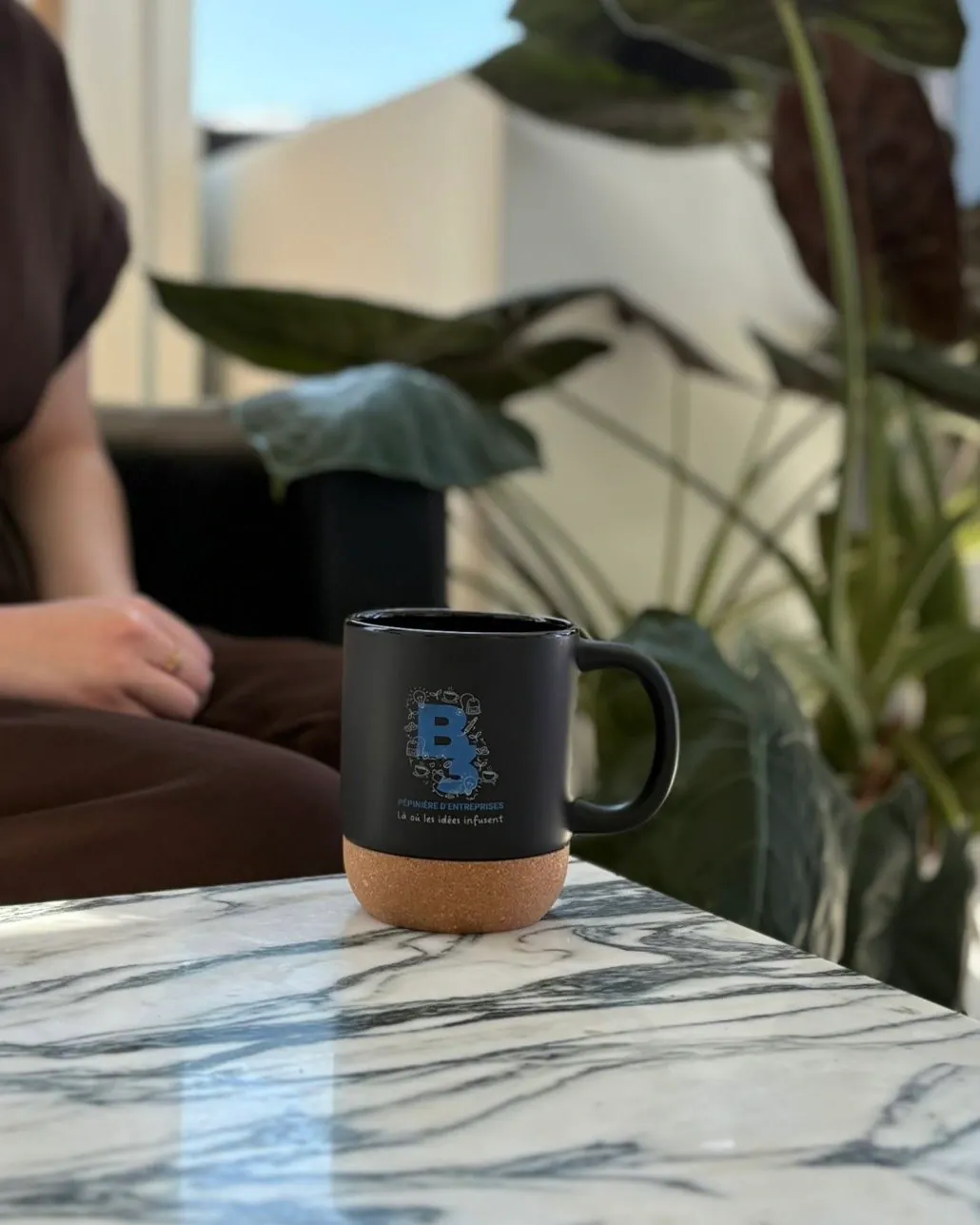La tasse noire à socle de liège posée sur une table en marbre, dans l’espace détente de la pépinière, une personne assise en arrière-plan