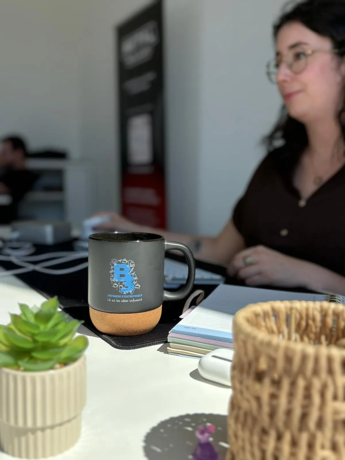 La tasse posée sur un bureau à côté d’un clavier et d’un carnet, une personne travaillant devant son écran en arrière-plan