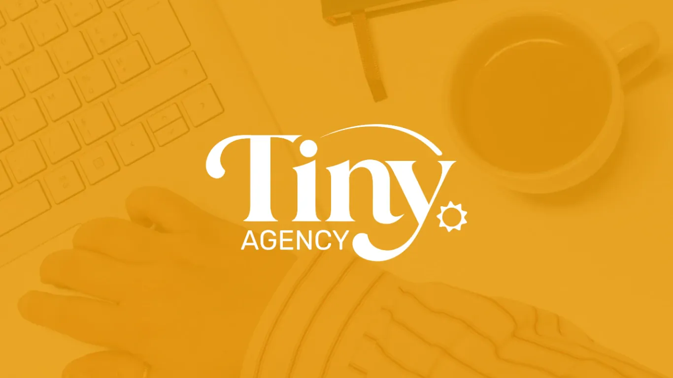Le logo Tiny Agency en blanc, « Tiny » en grandes lettres courbes surmonté d’un arc et suivi d’un petit soleil, « Agency » plus petit en dessous, sur une photo de bureau voilée d’orange : un clavier, un carnet, une tasse de thé et une main